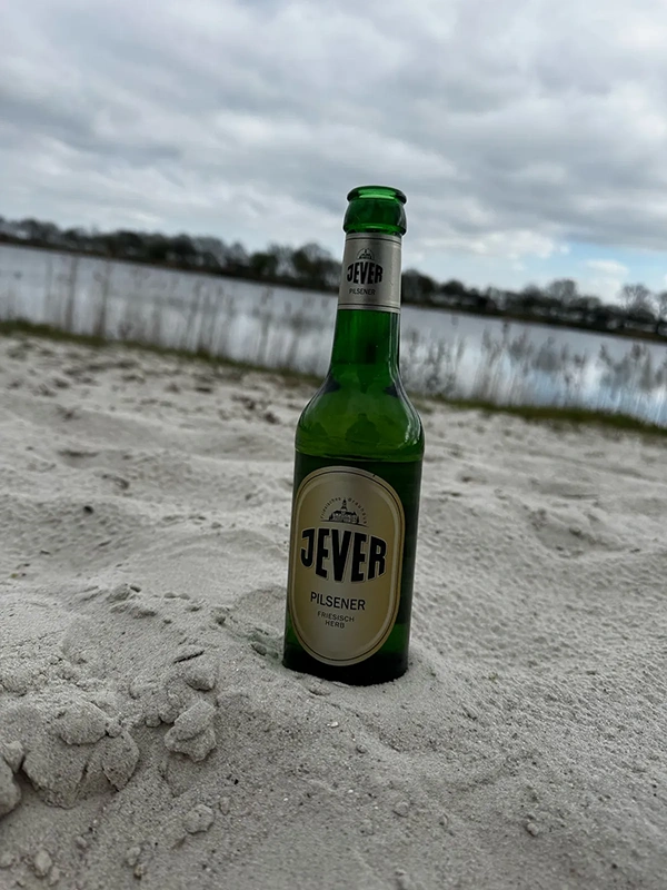 Eine grüne Bierflasche steht im Sand vor einem unscharfen Gewässer.