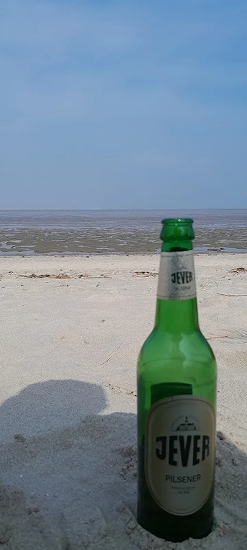 Eine grüne Bierflasche steht im Vordergrund auf einem Sandstrand mit Blick auf das Meer.