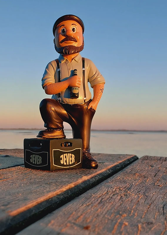 Eine Figur mit Bart und Hosenträgern hält eine Flasche und sitzt auf einem Koffer bei Sonnenuntergang.