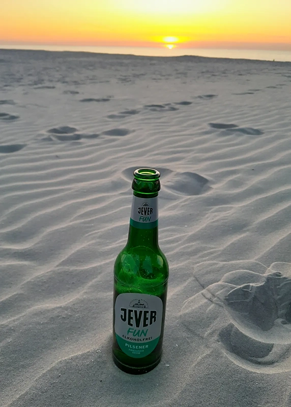 Grüne Flasche steht im Sand mit Sonnenuntergang im Hintergrund. Der Himmel färbt sich orange.