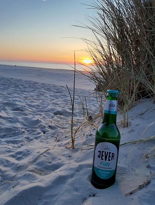 Eine Flasche steht im Vordergrund auf Sand mit Sonnenuntergang und Dünen im Hintergrund.