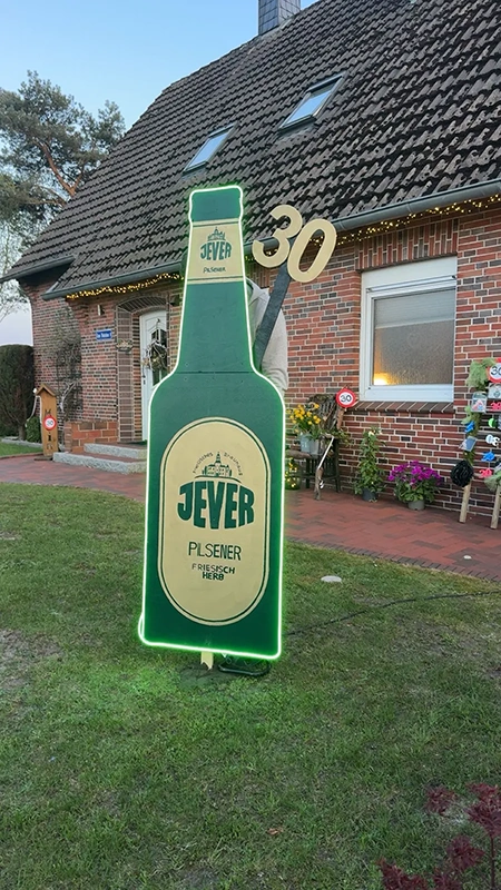 Eine beleuchtete Flaschenskulptur vor einem Backsteinhaus steht in einem Garten.