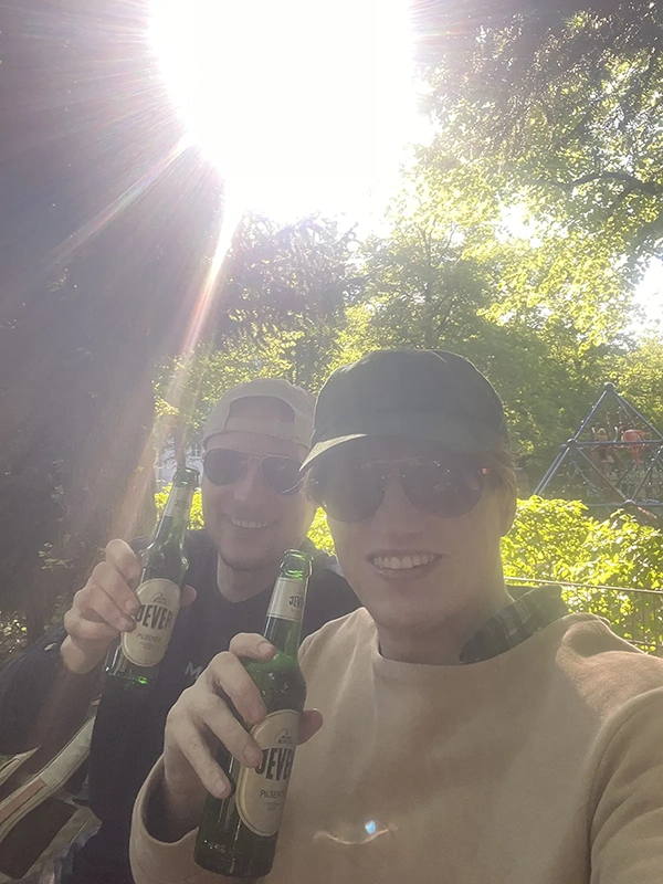 Zwei junge Männer lächeln und halten Flaschen in der Sonne. Sie tragen Sonnenbrillen und Baseballkappen.