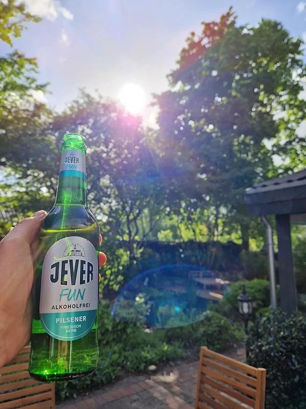 Eine Hand hält eine Flasche alkoholfreies Getränk in einem sonnigen Garten.
