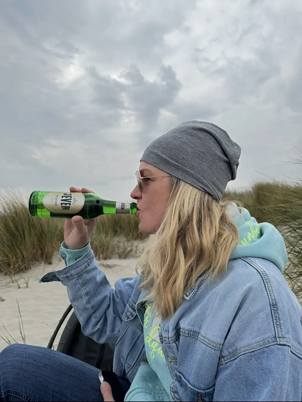 Eine Frau in einer Jeansjacke und Mütze trinkt aus einer Flasche in einer Dünenlandschaft.