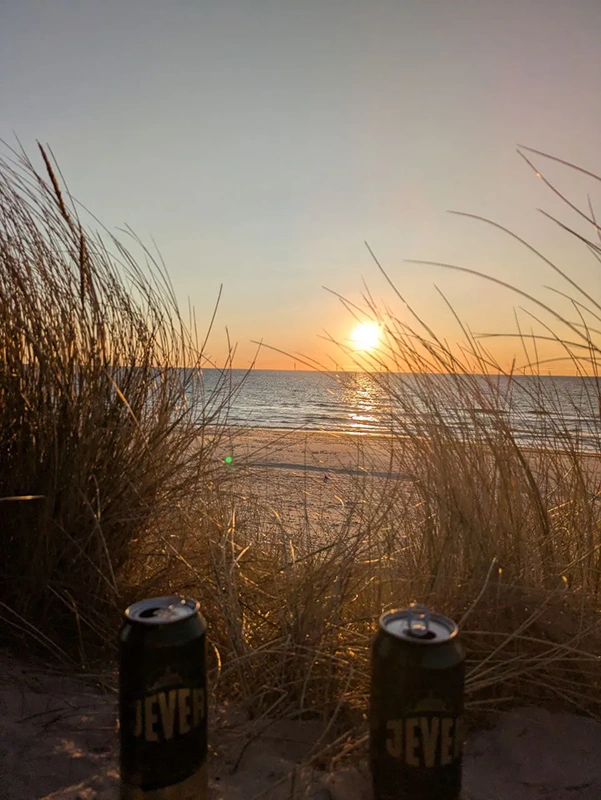 Sonnenuntergang am Strand mit zwei Getränkedosen im Vordergrund und Dünengras.