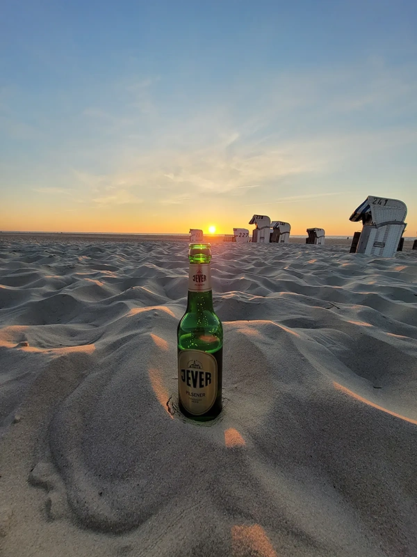 Eine grüne Flasche steht im Vordergrund auf sandigem Strand bei Sonnenuntergang. Im Hintergrund sind Strandkörbe zu erkennen.