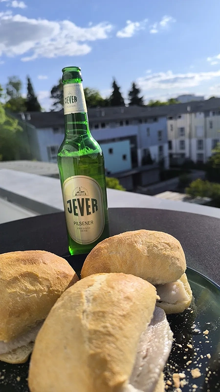 Eine grüne Bierflasche steht neben belegten Brötchen auf einem Tisch. Im Hintergrund sieht man Häuser und Bäume.
