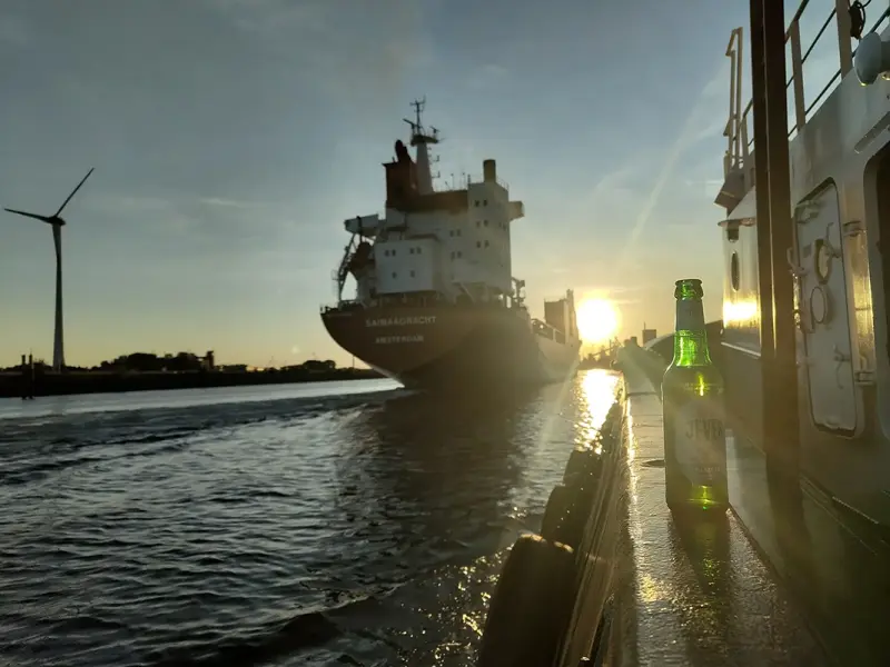 Eine Flasche Bier steht auf der Reling, während die Sonne hinter einem Schiff untergeht.