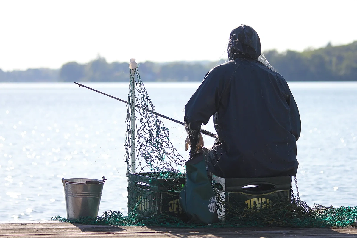 Eine Person in schwarzer Regenkleidung sitzt am Ufer und sortiert Angelnetze neben einem Eimer.