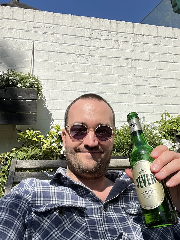 Ein Mann in Sonnenbrille sitzt lächelnd draußen und hält eine Flasche.