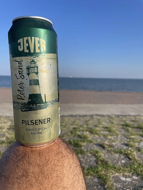 Eine Bierdose wird vor einer Strandkulisse in der Hand gehalten. Die Meereslinie ist in der Ferne sichtbar.