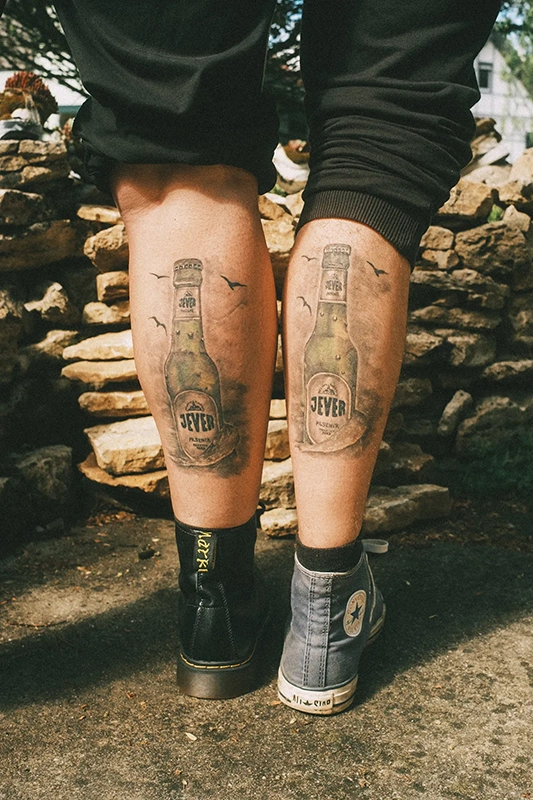 Zwei beinige Tätowierungen von Flaschen sind zwischen schwarzen Schuhen und blauen Schuhen sichtbar.