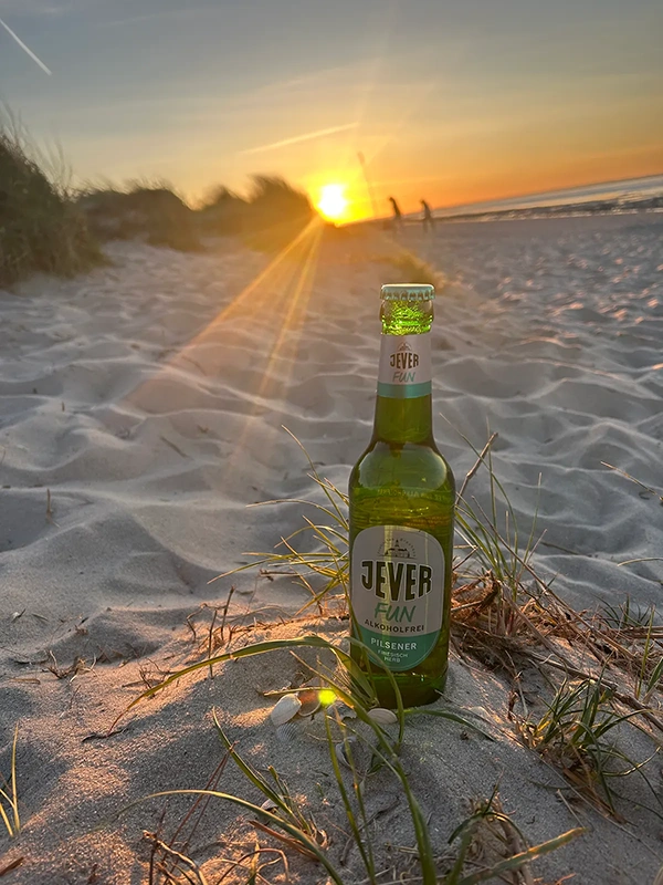 Jever Fun Flasche steht im Sand. Im Hintergrund Sonnenuntergang über dem Meer mit Dünen und Spaziergängern.