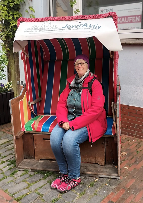 Eine Person sitzt auf einem bunt gestreiften Strandkorb. Im Hintergrund sind Pflastersteine und eine Hauswand.