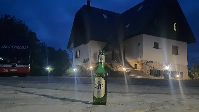 Eine Jever Pilsener Flasche steht auf gepflastertem Boden. Im Hintergrund ein beleuchtetes Wohnhaus bei Nacht.