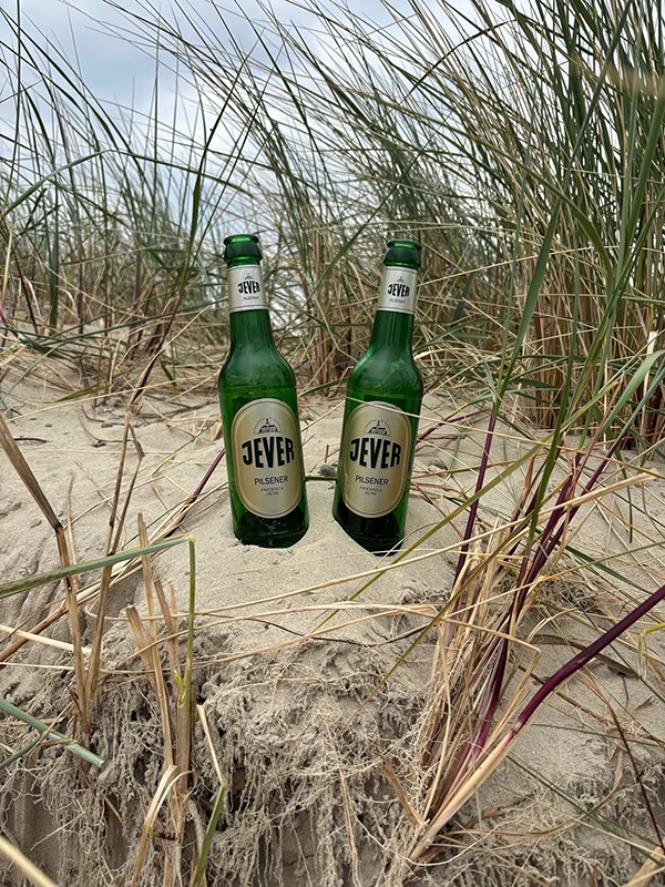 Zwei Jever Pilsener Flaschen stehen auf sandigem Boden. Im Hintergrund wachsen hohe Dünengräser.