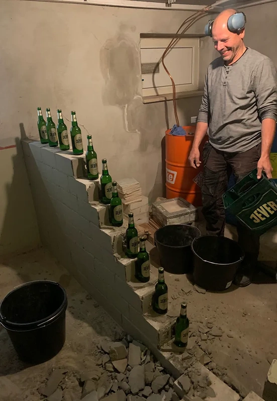 Eine gemauerte Treppe ist mit elf Jever Flaschen bestückt. Daneben steht ein lachender Mann am Arbeiten mit Gehörschutz.
