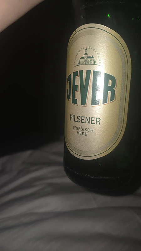 Jever Pilsener Flasche. Im Hintergrund ein dunkler Raum und ein helles Kissen.