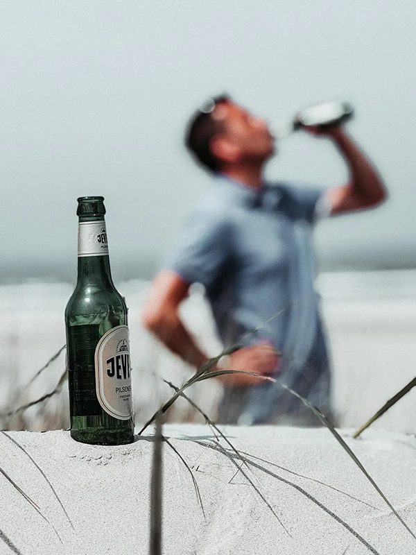 Jever Pilsener Flasche auf Sand in Nahaufnahme. Im Hintergrund unscharfer Mann am Strand.