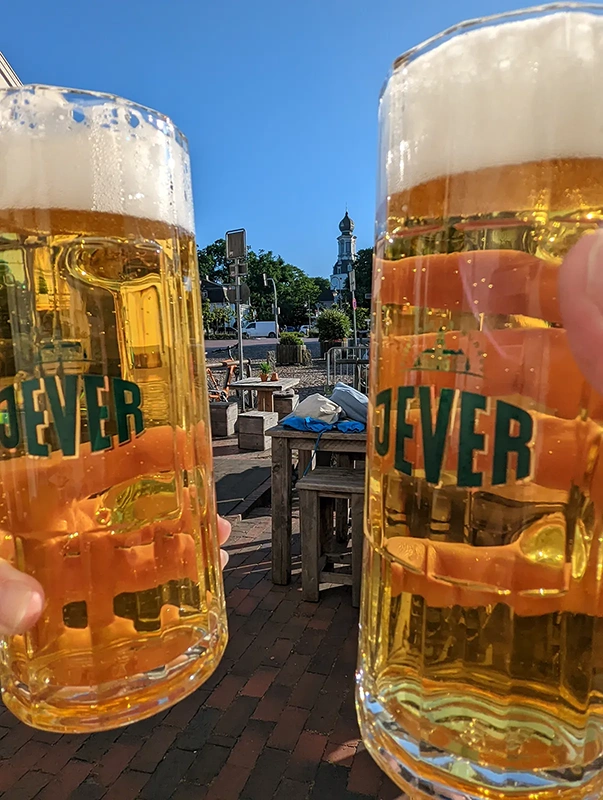 Zwei gefüllte Jever Biergläser werden angestoßen. Im Hintergrund stehen Tische auf einem Platz mit Kirchturm.