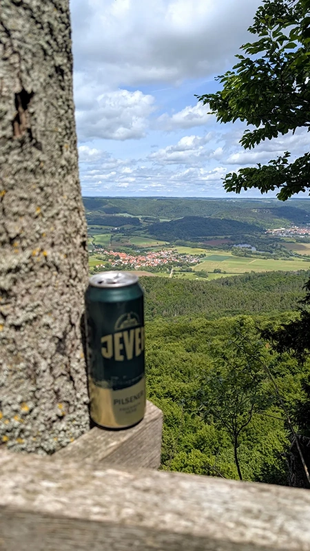 Eine Jever Pilsener Dose steht auf einem hölzernen Geländer neben einem Baumstamm. Im Hintergrund ein Tal mit Wäldern.