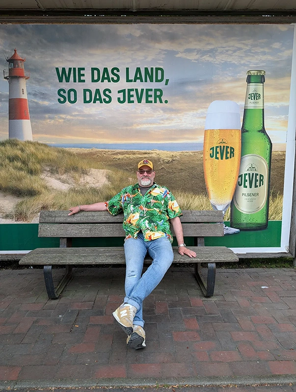 Ein Mann mit buntem Hawaiihemd sitzt auf einer Bank vor einem Werbeplakat mit Jever-Biermotiv.