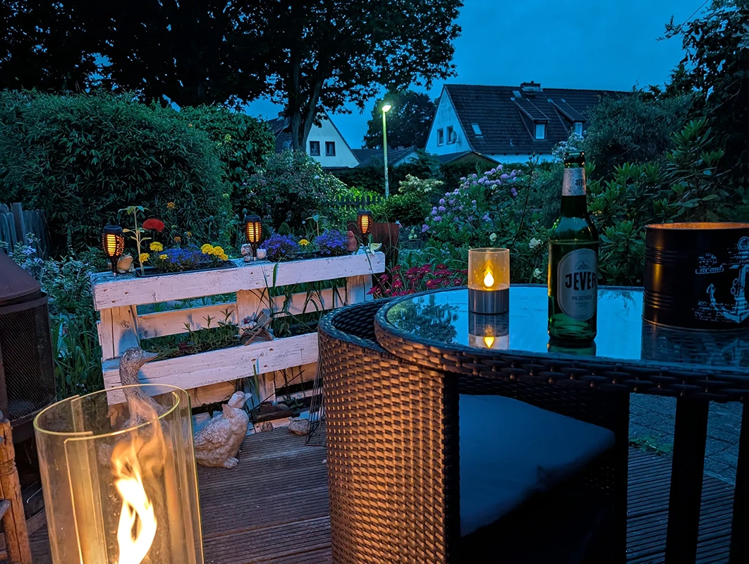 Terrassentisch mit Kerze und Jever Pilsener Flasche bei Abenddämmerung. Im Hintergrund Garten mit Blumen.