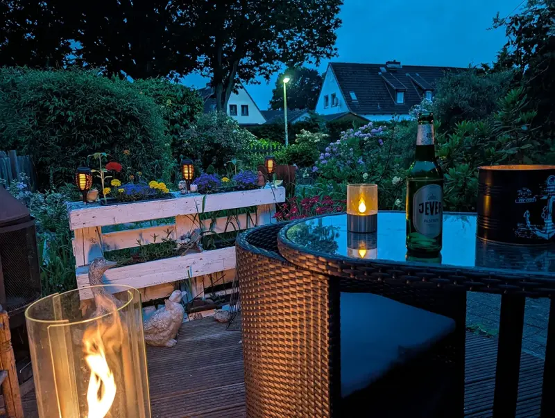Terrassentisch mit Kerze und Jever Pilsener Flasche bei Abenddämmerung. Im Hintergrund Garten mit Blumen.