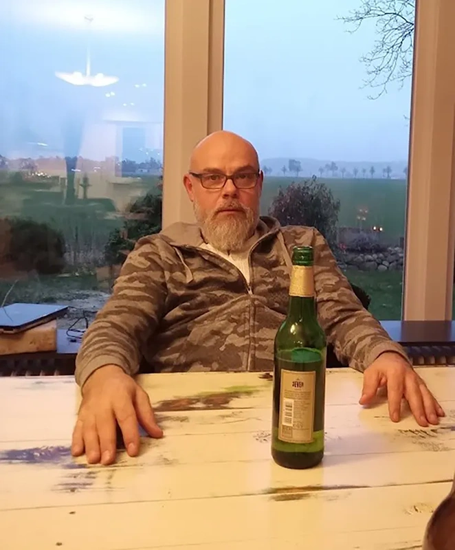 Mann mit Brille und Bart sitzt am Holztisch mit Jever Pilsener Flasche im Vordergrund, Landschaft  im Hintergrund.