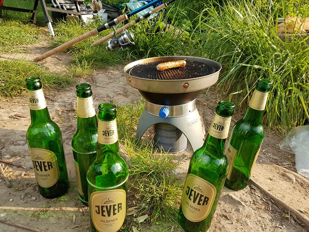 Fünf leere Jever Flaschen stehen um einen kleinen Grill mit einer Bratwurst. Im Hintergrund Gras und Angelausrüstung.