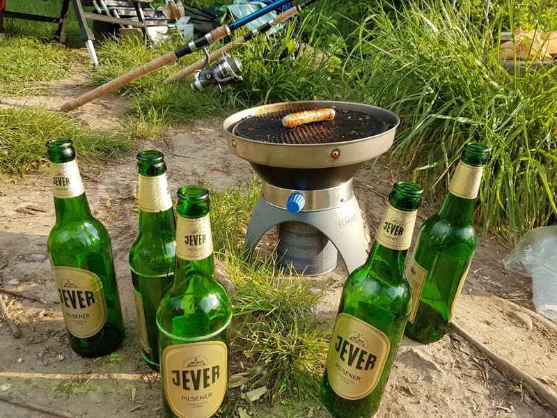 Fünf leere Jever Flaschen stehen um einen kleinen Grill mit einer Bratwurst. Im Hintergrund Gras und Angelausrüstung.