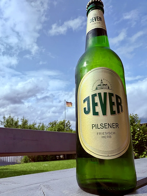 Jever-Pilsener Flasche auf einer Tischkante im Vordergrund. Im Hintergrund blauer Himmel mit Wolken.