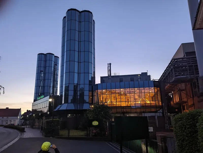Drei gläserne Türme mit Spiegelung, rechts beleuchtete Braukessel in verglastem Anbau, umgeben von Brauereianlagen.