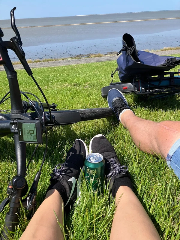 Zwei Personen sitzen mit Fahrrädern auf einer Wiese, eine Dose Jever zwischen den Füßen. Im Hintergrund ein Deich am Meer.