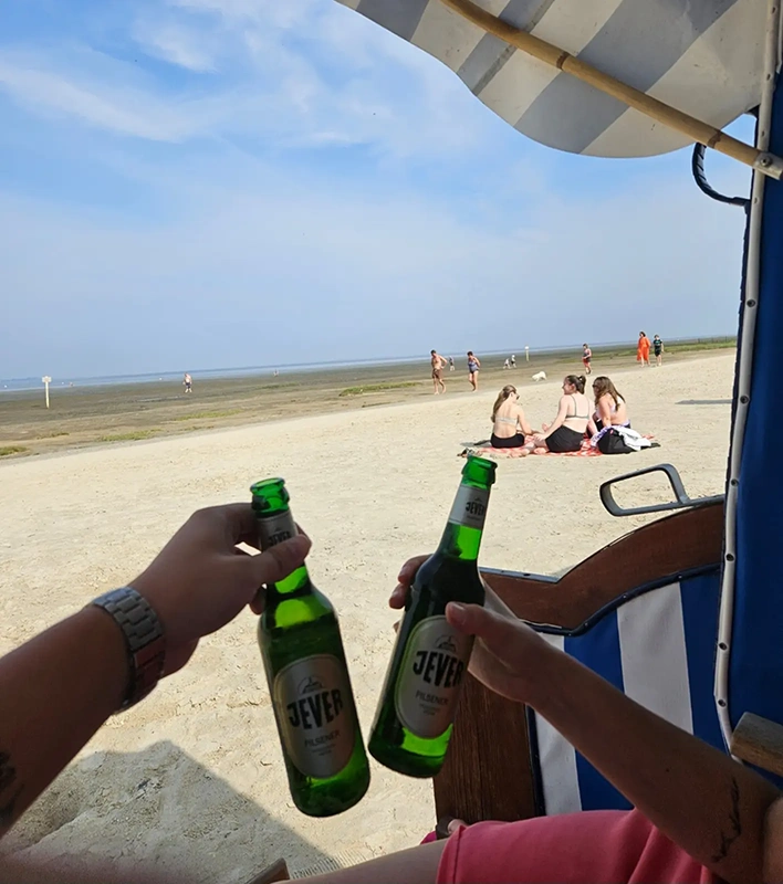 Zwei Hände stoßen mit Jever-Bierflaschen an, im Vordergrund ein Strandkorb. Im Hintergrund Menschen am Sandstrand.