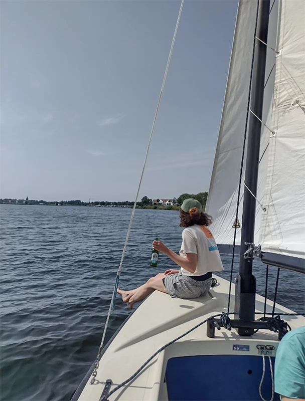 Eine Person sitzt auf einem Segelboot und hält eine Jever-Bierflasche in der Hand. Im Hintergrund Wasser und Häuser am Ufer.