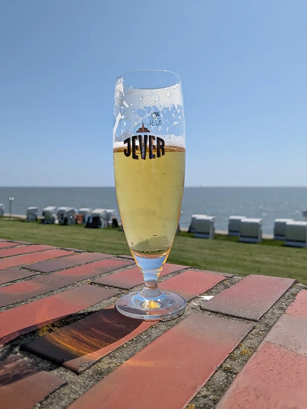 Ein gefülltes Jever-Glas steht auf einer roten Backsteinmauer. Im Hintergrund Strandkörbe und Meer unter blauem Himmel.