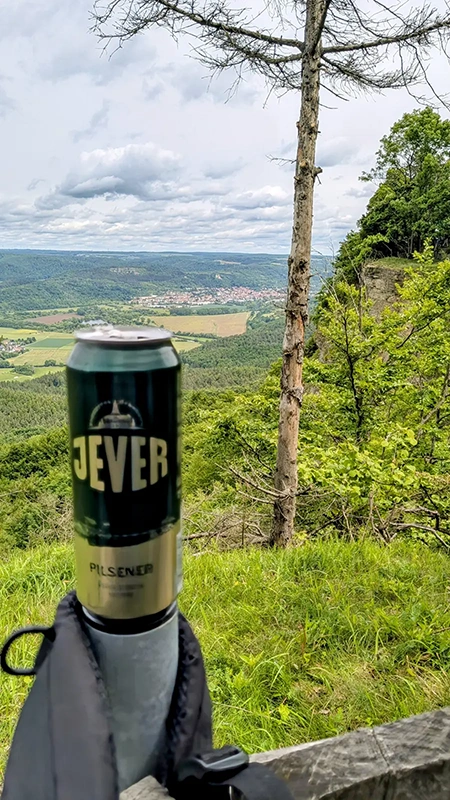 Eine Jever-Dose steht auf einem Geländer mit Blick auf ein bewaldetes Tal. Im Hintergrund Wiesen, Hügel und eine Ortschaft.