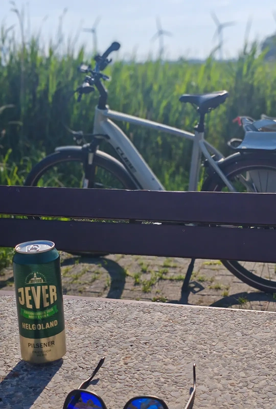 Eine Jever-Dose steht auf einem Steintisch neben einer Sonnenbrille. Im Hintergrund eine Bank, ein Fahrrad und ein Feld.