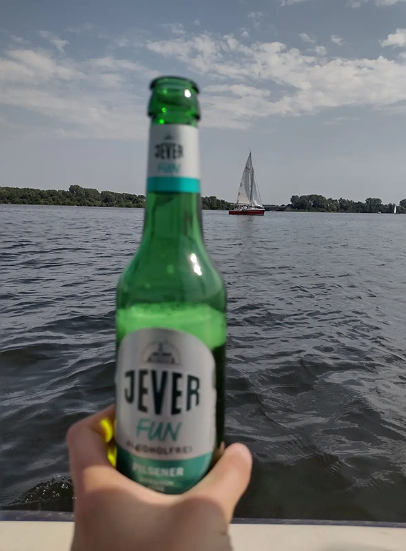 Eine Hand hält eine Jever-Flasche mit Blick auf das Wasser. Im Hintergrund fährt ein Segelboot vor einer Baumreihe entlang.
