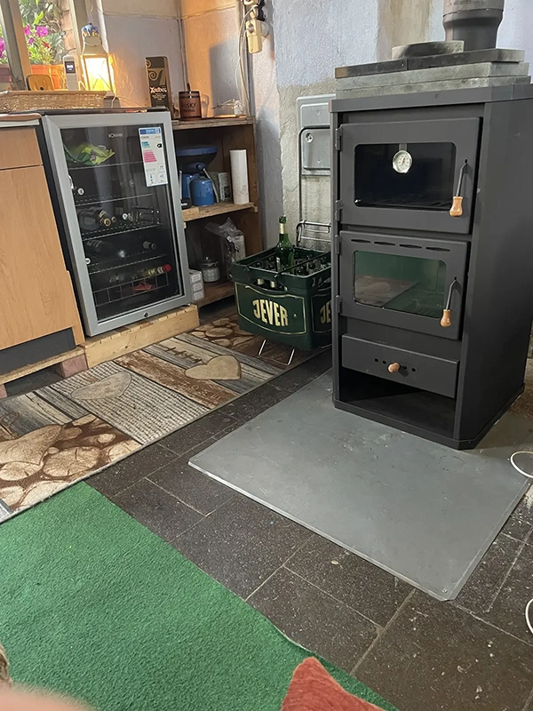 Ein Jever-Kasten steht neben einem schwarzen Ofen. Im Hintergrund ein Kühlschrank und Regale mit Haushaltsgegenständen.