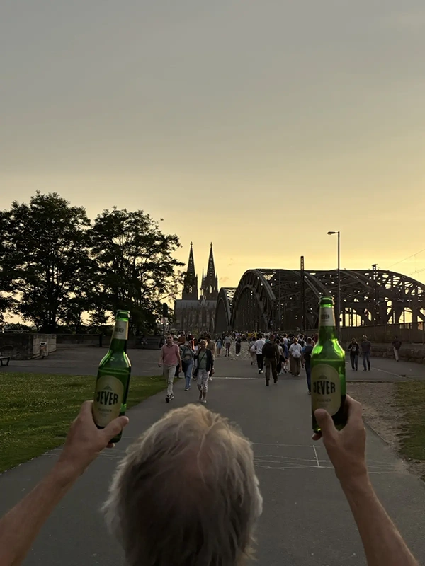 Eine Person hält zwei Jever-Flaschen in die Luft. Im Hintergrund Hohenzollernbrücke, Kölner Dom und Menschen im Abendlicht.