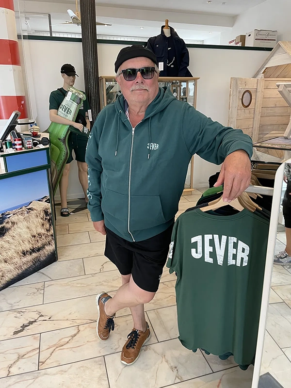 Älterer Mann mit Sonnenbrille, Mütze und Jever-Kapuzenjacke steht in einem Jever Shop neben einem Jever-T-Shirt.	