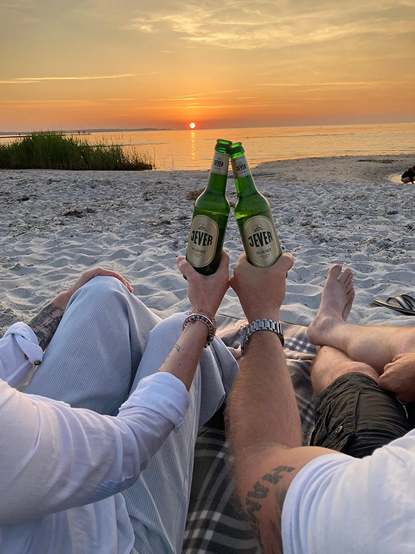 Zwei Personen stoßen mit Jever-Bierflaschen an. Im Hintergrund Sonnenuntergang über dem Meer mit Strand und Gras.