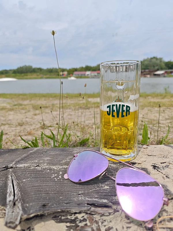 Ein Jever Bierglas steht auf einem Baumstamm neben einer Sonnenbrille mit lila Gläsern. Im Hintergrund ist ein See.