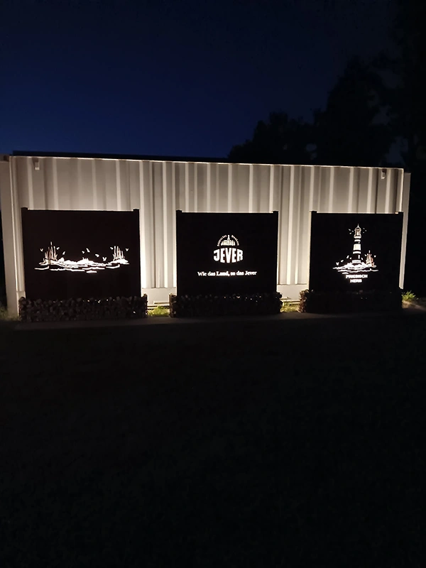 Drei beleuchtete Metalltafeln mit Jever Motiven und Schriftzug Jever stehen vor einer weißen Containerwand bei Nacht.