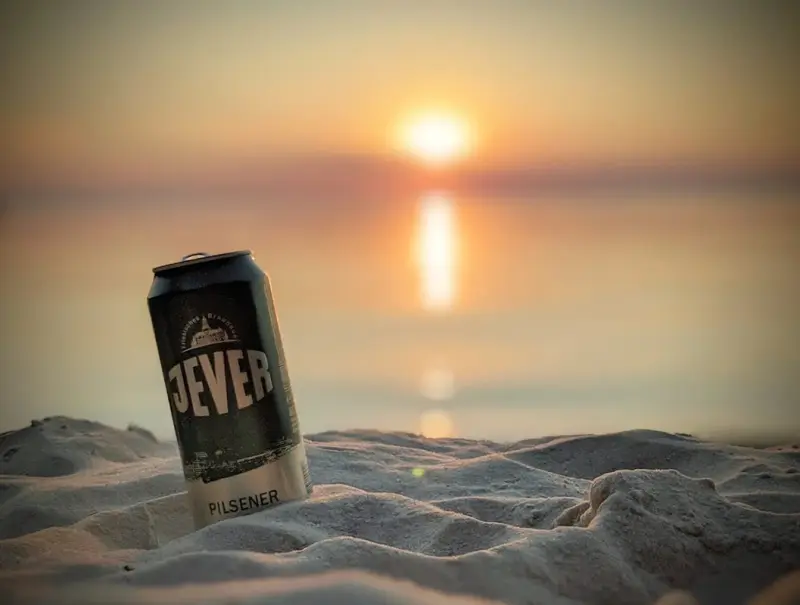 Eine Dose Jever Pilsener steht auf hellem Sand. Im Hintergrund ist ein Sonnenuntergang über dem Meer zu sehen.