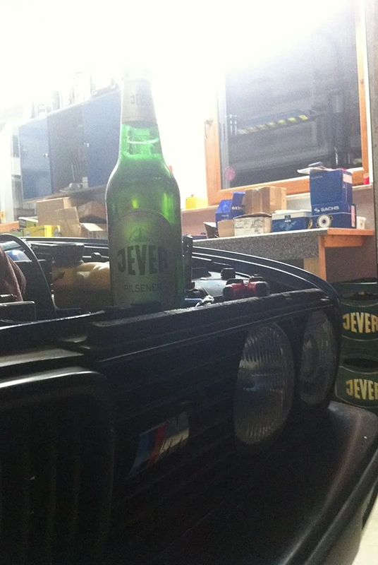 Jever Bierflasche steht auf dem Kühlergrill eines schwarzen BMW mit Doppelscheinwerfern. Im Hintergrund Werkstattregale.