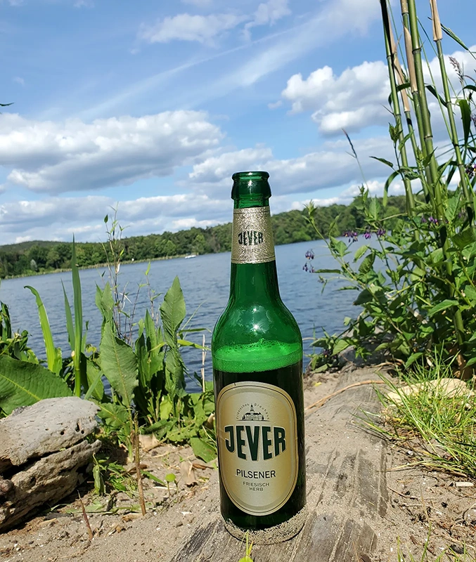 Jever Bierflasche steht auf sandigem Boden. Im Hintergrund ein See mit Pflanzen und leicht bewölktem Himmel.